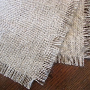 May include: Two pieces of natural burlap fabric with frayed edges. The fabric is a light beige color and has a woven texture.