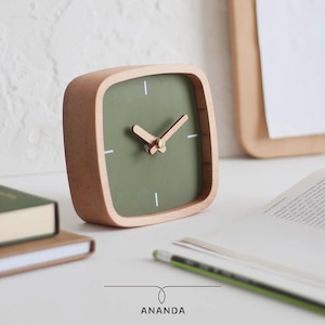 May include: A square wooden desk clock with a green face and light wood frame. The clock has white markers and light wood hands. The word "ANANDA" is printed at the bottom of the clock.