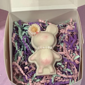 Może przedstawiać: Biały cukierek w kształcie misia z różowymi akcentami, perłowym naszyjnikiem i kwiatkiem na głowie, zapakowany w białe pudełko z fioletowym, różowym i miętowym wypełnieniem z papieru. Pudełko jest otwarte, ukazując zawartość.