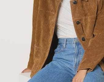 May include: A brown suede jacket with a collared neckline and button closure. The jacket is paired with a white turtleneck and blue denim jeans. The model is wearing silver hoop earrings.