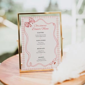 Op de afbeelding: Een kerstdiner menukaart in een goudkleurige metalen lijst, met een roze gingham achtergrond, een decoratieve rode lintrand en een roze strikillustratie. De tekst vermeldt voorgerechten, hoofdgerechten, drankjes en desserts voor het diner.