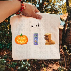 May include: A white t-shirt featuring the word mood in black script text above three graphics: an orange jack-o'-lantern pumpkin, a purple Alani energy drink can, and a tan suede winter boot with a brown ribbon bow on the side.