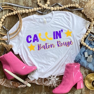 May include: A white t-shirt with the text "CA'LIN' Baton Rouge" in purple and yellow. The image also features a pair of pink ankle boots, denim jeans, and decorative beads.