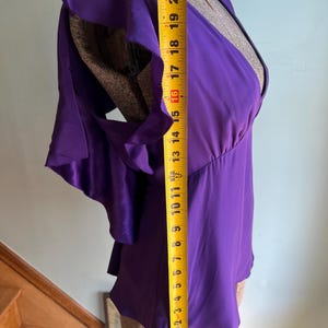 May include: A vibrant purple blouse with ruffled sleeves, measured at approximately 21 inches in length. The top features a V-neck design and is displayed on a mannequin. The fabric appears to be a smooth, silky material.