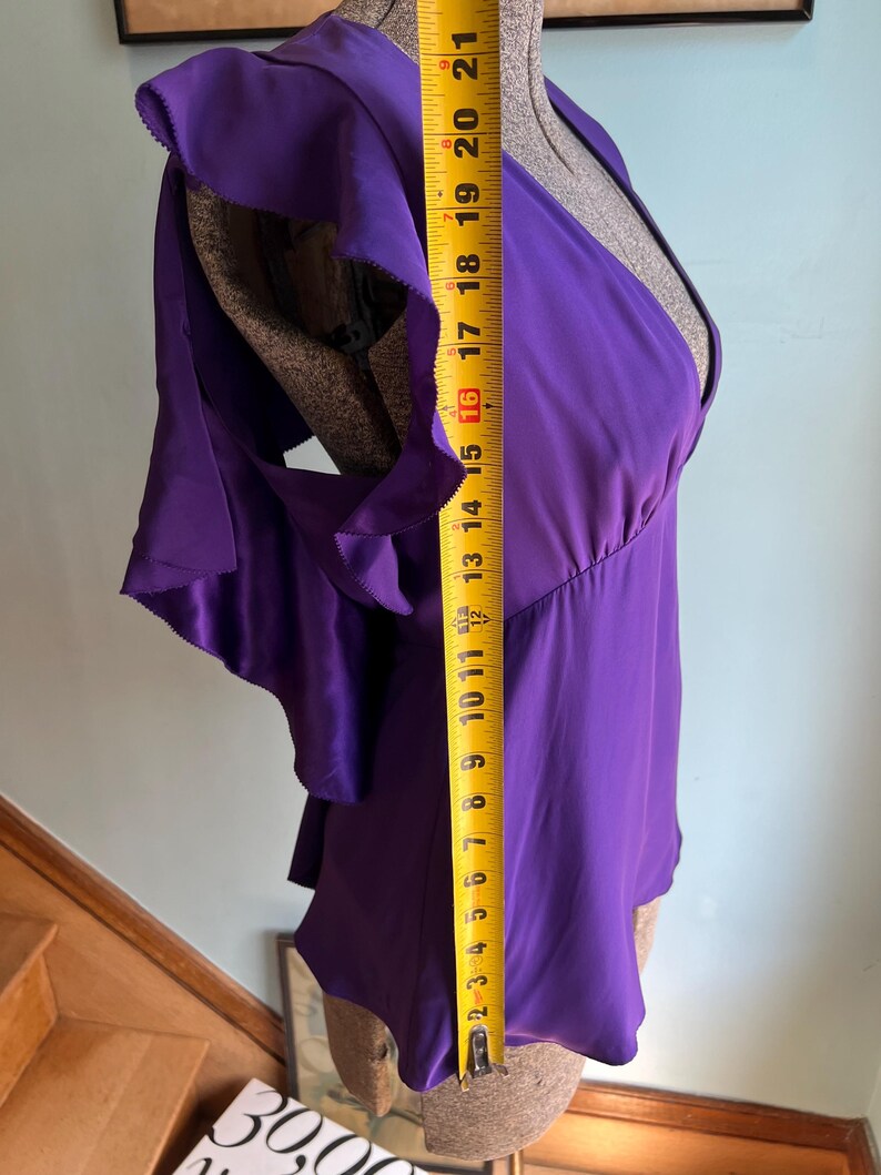 May include: A vibrant purple blouse with ruffled sleeves, measured at approximately 21 inches in length. The top features a V-neck design and is displayed on a mannequin. The fabric appears to be a smooth, silky material.