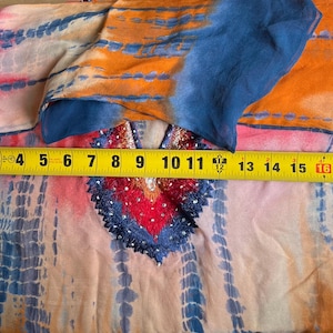 Peut inclure: Un v&ecirc;tement color&eacute; &agrave; motifs tie-dye avec un d&eacute;collet&eacute; d&eacute;coratif. Le tissu est principalement orange, bleu et rose, avec un m&egrave;tre ruban indiquant une largeur d'environ 46 cm.