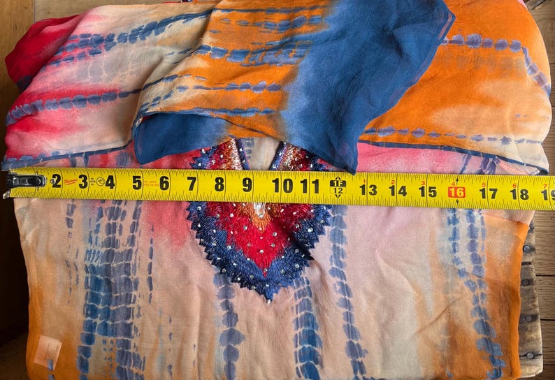 Peut inclure: Un v&ecirc;tement color&eacute; &agrave; motifs tie-dye avec un d&eacute;collet&eacute; d&eacute;coratif. Le tissu est principalement orange, bleu et rose, avec un m&egrave;tre ruban indiquant une largeur d'environ 46 cm.