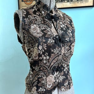 May include: A sleeveless vest featuring a black and brown floral brocade pattern with a high mandarin collar and three black decorative frog closures down the front. The garment is displayed on a mannequin, showcasing the intricate woven fabric.