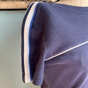 K&ouml;nnte beinhalten: Marineblaues T-Shirt mit wei&szlig;en und blauen Streifen am &Auml;rmel und einem wei&szlig;en diagonalen Streifen &uuml;ber der Brust. Das Shirt wird auf einer Schaufensterpuppe pr&auml;sentiert.