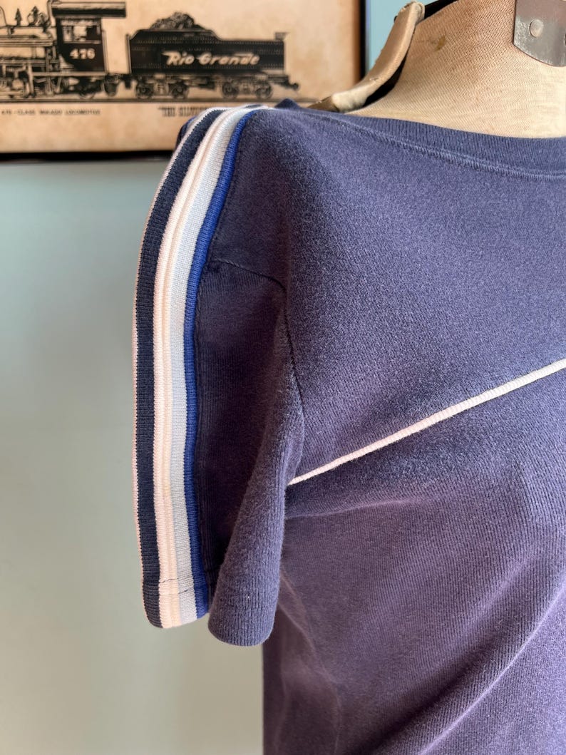 K&ouml;nnte beinhalten: Marineblaues T-Shirt mit wei&szlig;en und blauen Streifen am &Auml;rmel und einem wei&szlig;en diagonalen Streifen &uuml;ber der Brust. Das Shirt wird auf einer Schaufensterpuppe pr&auml;sentiert.