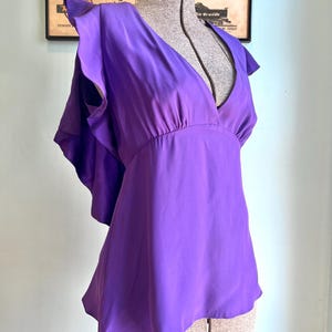 May include: A vibrant purple blouse with a deep V-neck and flutter sleeves. The top is made of a silky material and has a gathered waist. The blouse is displayed on a mannequin, showcasing its flowing design and rich color.