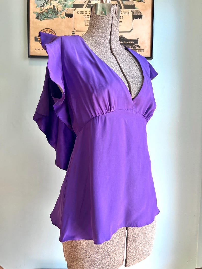 May include: A vibrant purple blouse with a deep V-neck and flutter sleeves. The top is made of a silky material and has a gathered waist. The blouse is displayed on a mannequin, showcasing its flowing design and rich color.