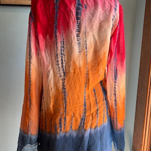 Peut inclure: Une chemise tie-dye &agrave; manches longues dans les tons de rouge, d'orange et de bleu. La chemise a un col en V et une coupe ample. Le motif tie-dye pr&eacute;sente des rayures verticales bleu fonc&eacute; sur le fond color&eacute;. La chemise est en tissu l&eacute;ger et transparent.