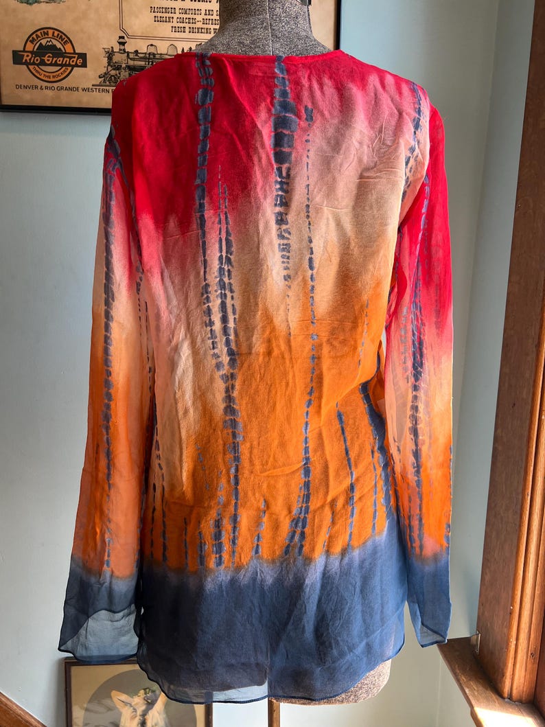Peut inclure: Une chemise tie-dye &agrave; manches longues dans les tons de rouge, d'orange et de bleu. La chemise a un col en V et une coupe ample. Le motif tie-dye pr&eacute;sente des rayures verticales bleu fonc&eacute; sur le fond color&eacute;. La chemise est en tissu l&eacute;ger et transparent.
