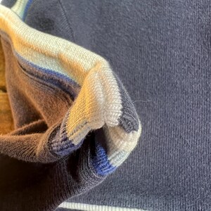 K&ouml;nnte beinhalten: Nahaufnahme eines marineblauen Pullovers mit wei&szlig;en und blauen Streifen am &Auml;rmel. Das &Auml;rmelb&uuml;ndchen hat ein Streifendesign in Wei&szlig;, Hellblau und Dunkelblau. Der Pullover hat einen wei&szlig;en Streifen am unteren Saum.