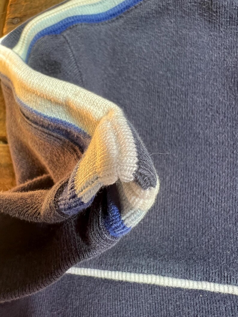 K&ouml;nnte beinhalten: Nahaufnahme eines marineblauen Pullovers mit wei&szlig;en und blauen Streifen am &Auml;rmel. Das &Auml;rmelb&uuml;ndchen hat ein Streifendesign in Wei&szlig;, Hellblau und Dunkelblau. Der Pullover hat einen wei&szlig;en Streifen am unteren Saum.
