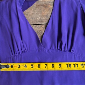 May include: A vibrant purple dress with a deep V-neck and flutter sleeves. The dress is measured with a yellow tape measure, showing a width of approximately 12 inches across the bodice. The fabric appears to be a lightweight, flowing material.