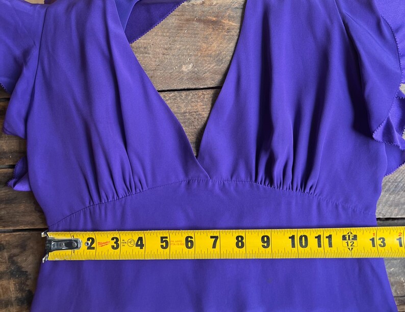 May include: A vibrant purple dress with a deep V-neck and flutter sleeves. The dress is measured with a yellow tape measure, showing a width of approximately 12 inches across the bodice. The fabric appears to be a lightweight, flowing material.