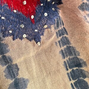 Peut inclure: Gros plan sur un textile avec des d&eacute;tails brod&eacute;s. Le motif pr&eacute;sente une zone centrale de broderie rouge et bleue, orn&eacute;e de petites perles argent&eacute;es. Le tissu a une base beige avec des motifs bleus de style tie-dye sur les bords.