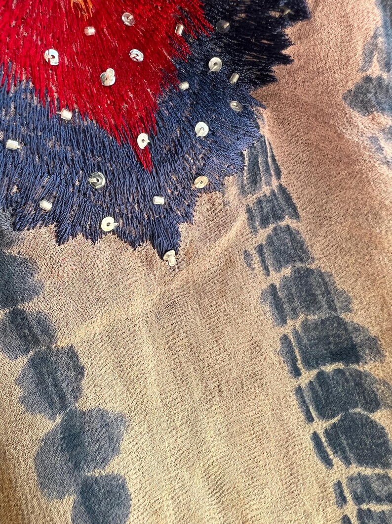 Peut inclure: Gros plan sur un textile avec des d&eacute;tails brod&eacute;s. Le motif pr&eacute;sente une zone centrale de broderie rouge et bleue, orn&eacute;e de petites perles argent&eacute;es. Le tissu a une base beige avec des motifs bleus de style tie-dye sur les bords.