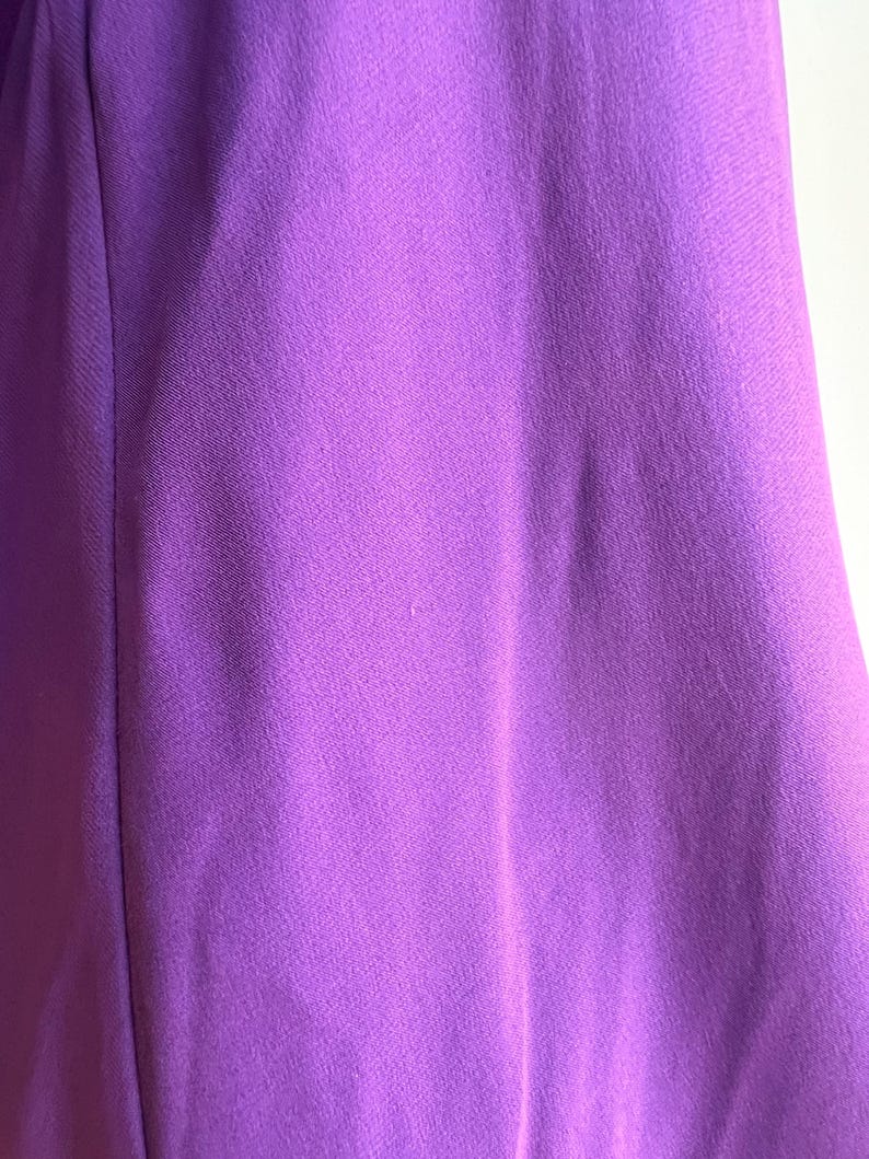 May include: Close-up of a vibrant purple fabric. The material has a subtle texture and a smooth appearance. The color is a rich, saturated shade of purple, with slight variations in tone across the surface. The fabric appears to be a solid color.