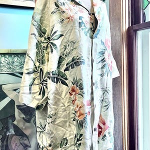 May include: A long-sleeved, button-down shirt with a floral pattern in shades of pink, orange, and green on a cream background. The shirt is displayed on a mannequin. The shirt has a collar and buttons down the front.
