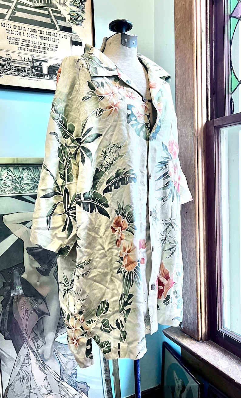 May include: A long-sleeved, button-down shirt with a floral pattern in shades of pink, orange, and green on a cream background. The shirt is displayed on a mannequin. The shirt has a collar and buttons down the front.