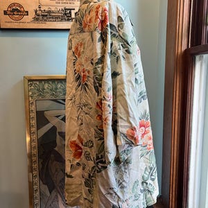 May include: A cream-colored, long-sleeved shirt with a vibrant floral pattern in shades of red, orange, and green. The shirt is displayed on a mannequin. The shirt has a collar and buttoned cuffs. The background includes a vintage travel poster.