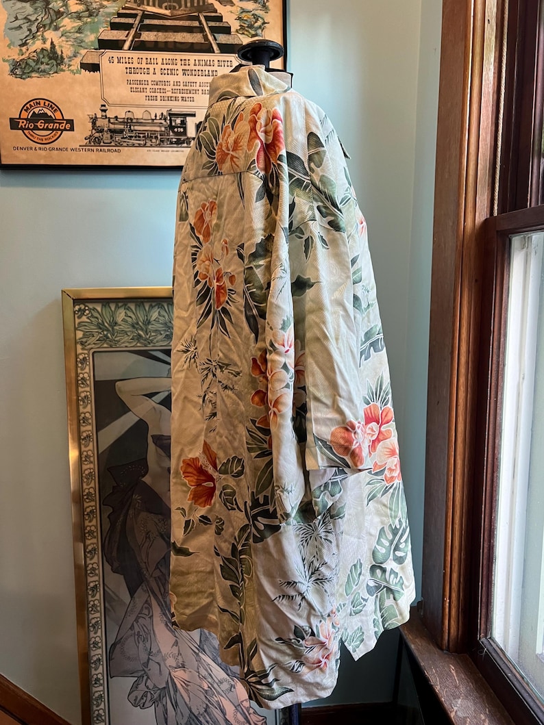 May include: A cream-colored, long-sleeved shirt with a vibrant floral pattern in shades of red, orange, and green. The shirt is displayed on a mannequin. The shirt has a collar and buttoned cuffs. The background includes a vintage travel poster.