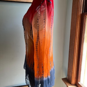 Peut inclure: Une tunique &agrave; manches longues, tie-dye, dans les tons de rouge, d'orange et de bleu. Le tissu transparent est ample, avec un effet d&eacute;grad&eacute; du rouge aux &eacute;paules au bleu &agrave; l'ourlet. La tunique est pr&eacute;sent&eacute;e sur un mannequin.