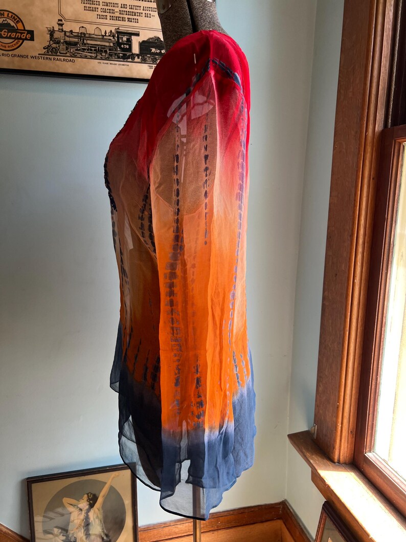 Peut inclure: Une tunique &agrave; manches longues, tie-dye, dans les tons de rouge, d'orange et de bleu. Le tissu transparent est ample, avec un effet d&eacute;grad&eacute; du rouge aux &eacute;paules au bleu &agrave; l'ourlet. La tunique est pr&eacute;sent&eacute;e sur un mannequin.