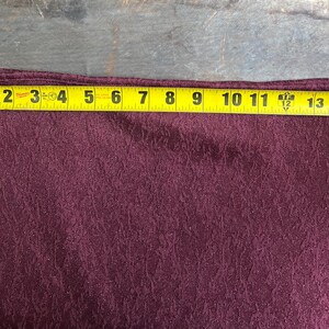 May include: A burgundy fabric with a textured pattern. The fabric is measured with a tape measure, showing a length of 15 inches.