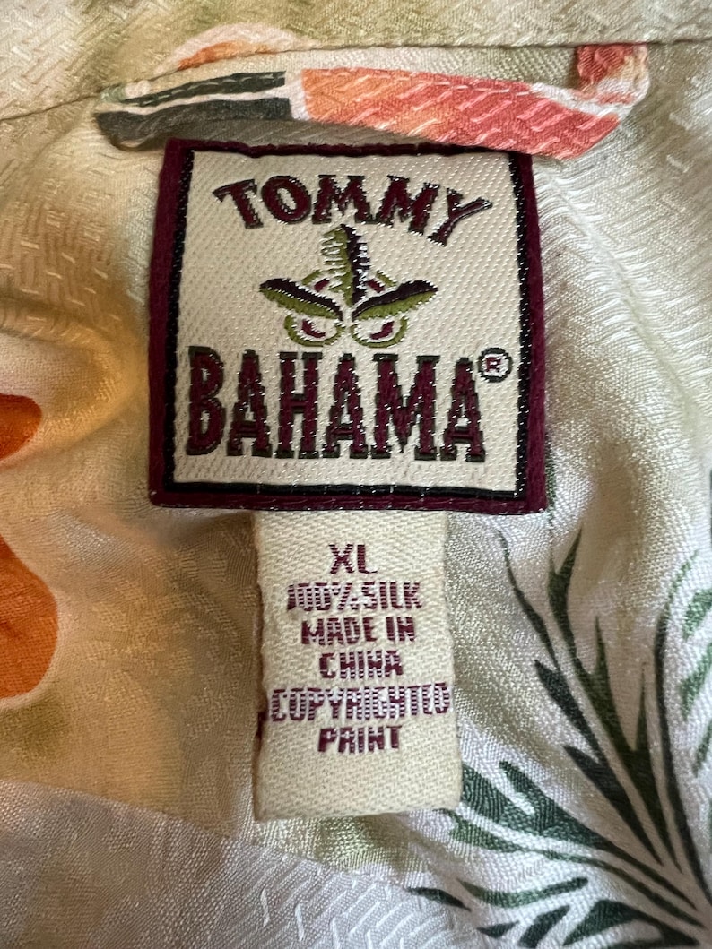 May include: Close-up of a Tommy Bahama shirt tag. The tag is rectangular with a maroon border and the brand name in maroon. The tag reads "XL 100% Silk Made in China Copyrighted Print."