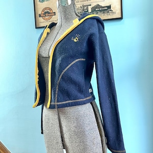 May include: A navy blue fleece zip-up hoodie featuring a bright yellow trim along the zipper and hood edge. The jacket includes long sleeves, a small yellow handprint graphic on the upper left chest, and visible yellow stitching along the seams.