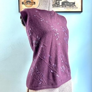 May include: A sleeveless plum-colored knit top featuring a crew neckline and delicate tonal embroidery of winding floral vines and leaves across the front. The garment is displayed on a mannequin, showcasing its soft texture and straight hemline.
