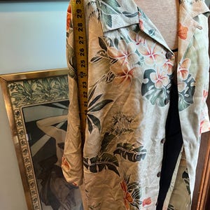 May include: A beige button-down shirt with a tropical floral print in orange, green, and white. The shirt is measured at approximately 30 inches in length. The shirt is displayed on a mannequin.