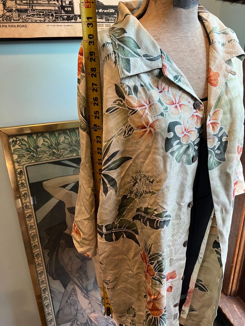 May include: A beige button-down shirt with a tropical floral print in orange, green, and white. The shirt is measured at approximately 30 inches in length. The shirt is displayed on a mannequin.