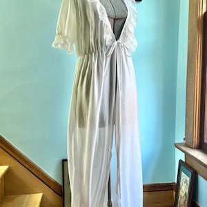 May include: A floor-length white robe featuring short sleeves with ruffled lace trim, a deep V-neckline edged in matching white lace, and a thin fabric tie closure at the waist. The garment is made of a lightweight, semi-sheer fabric.