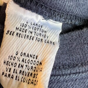K&ouml;nnte beinhalten: Nahaufnahme eines wei&szlig;en Kleidungsetiketts mit schwarzem Text. Das Etikett lautet "L LARGE 100% COTTON MADE IN TURKEY SEE REVERSE FOR CARE". Das Etikett ist an einem dunkelblauen Stoff befestigt.