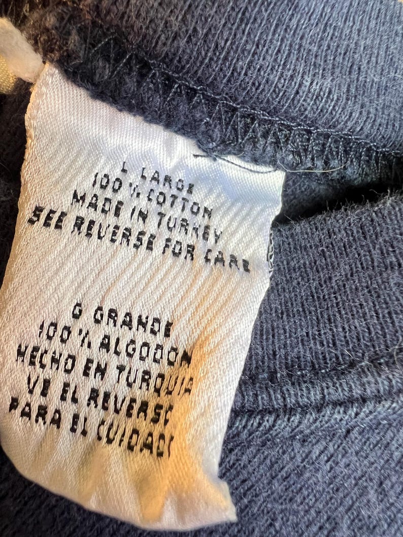 K&ouml;nnte beinhalten: Nahaufnahme eines wei&szlig;en Kleidungsetiketts mit schwarzem Text. Das Etikett lautet "L LARGE 100% COTTON MADE IN TURKEY SEE REVERSE FOR CARE". Das Etikett ist an einem dunkelblauen Stoff befestigt.