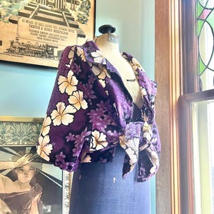 May include: A vintage purple jacket with a floral pattern of white and yellow hibiscus flowers and smaller purple flowers. The jacket is displayed on a mannequin with a blue dress form. The jacket has a tie closure and button details.