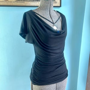 May include: A black short-sleeved top featuring a draped cowl neckline and gathered side seams. The garment is displayed on a textured gray mannequin torso and includes a thin black cord necklace with a circular silver-toned pendant at the center.