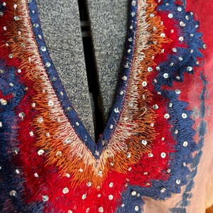 Peut inclure: Gros plan sur un v&ecirc;tement color&eacute; avec un col en V. Le col est orn&eacute; de broderies complexes dans des tons de rouge, d'orange et de bleu, rehauss&eacute;es de petits sequins scintillants. Le tissu pr&eacute;sente un effet tie-dye avec des nuances de rouge, de bleu et de p&ecirc;che.