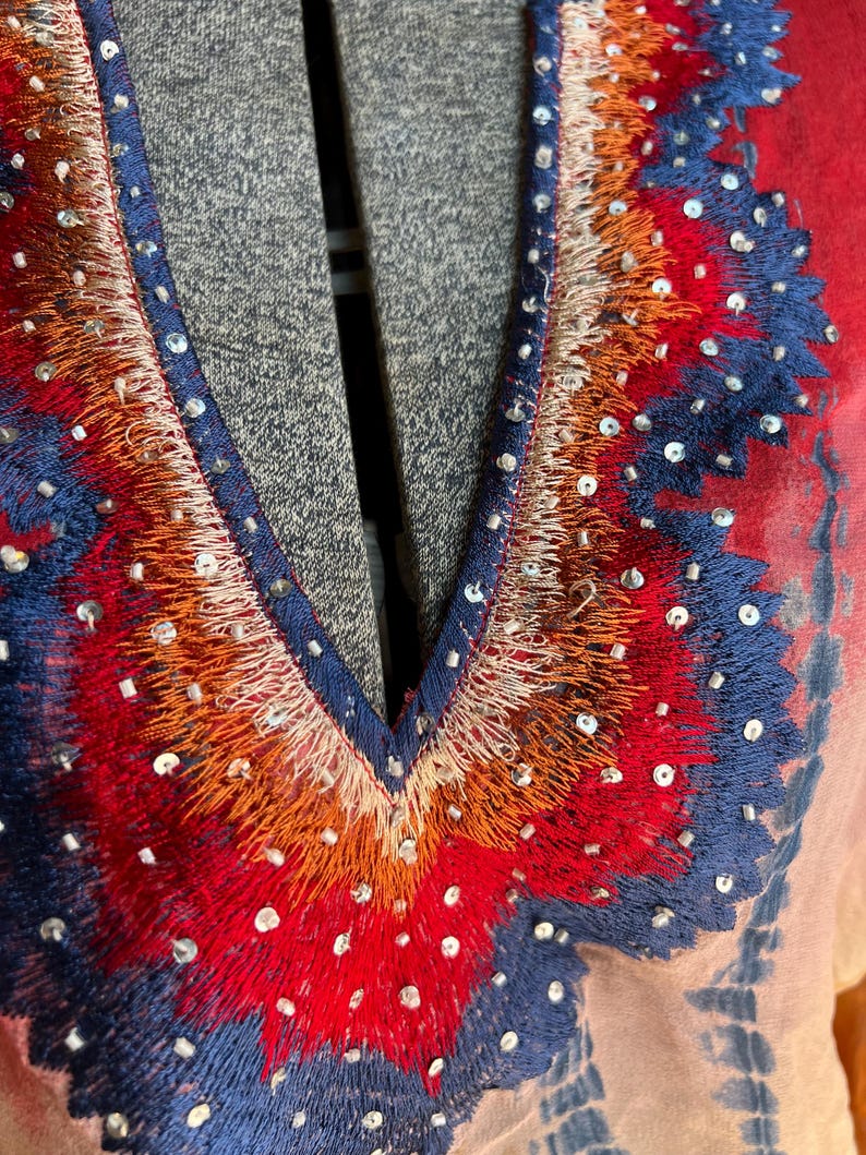 Peut inclure: Gros plan sur un v&ecirc;tement color&eacute; avec un col en V. Le col est orn&eacute; de broderies complexes dans des tons de rouge, d'orange et de bleu, rehauss&eacute;es de petits sequins scintillants. Le tissu pr&eacute;sente un effet tie-dye avec des nuances de rouge, de bleu et de p&ecirc;che.