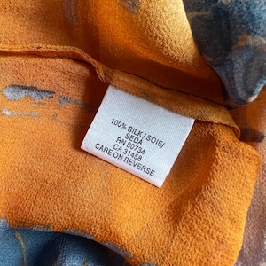 Peut inclure: Gros plan sur un tissu de soie avec un motif abstrait orange et bleu. Une &eacute;tiquette blanche indique "100% SILK/SOIE/SEDA" et "CARE ON REVERSE". Le tissu a une texture douce et fluide.