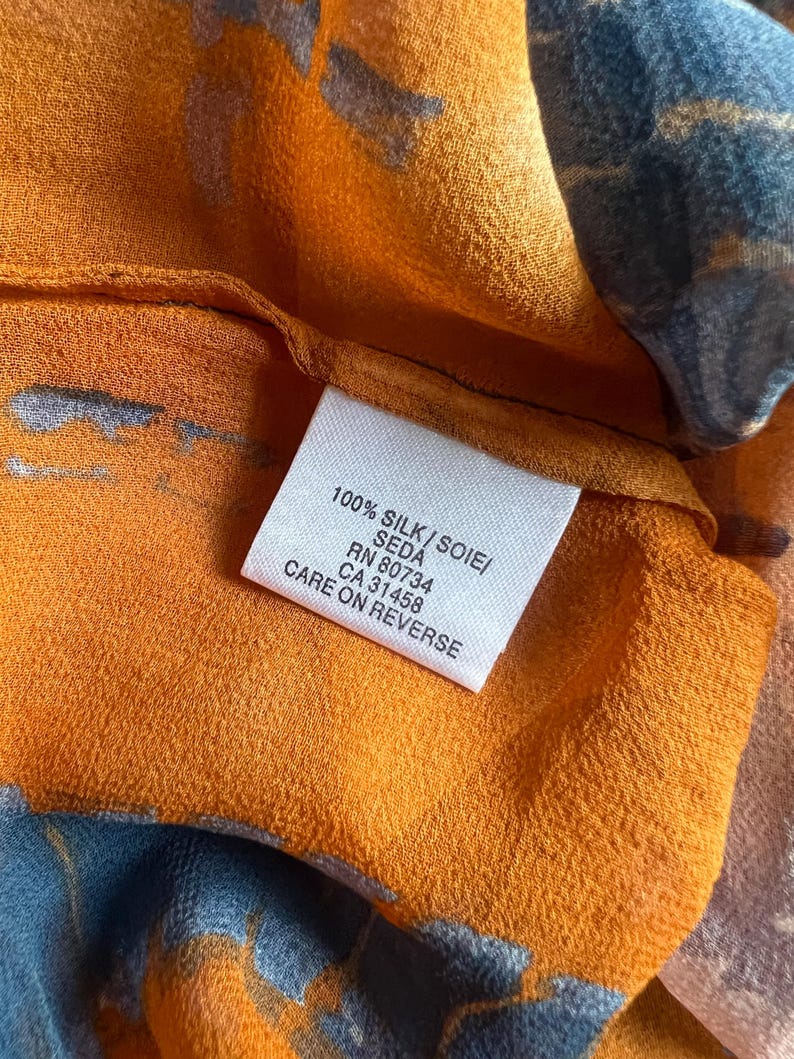 Peut inclure: Gros plan sur un tissu de soie avec un motif abstrait orange et bleu. Une &eacute;tiquette blanche indique "100% SILK/SOIE/SEDA" et "CARE ON REVERSE". Le tissu a une texture douce et fluide.