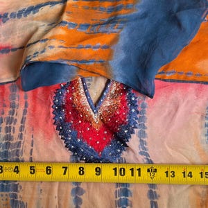 Peut inclure: Une tunique color&eacute;e de style tie-dye avec un d&eacute;collet&eacute; en V. La tunique pr&eacute;sente un m&eacute;lange vibrant de teintes orange, rose et bleu. L'encolure est orn&eacute;e de broderies et de sequins. Le m&egrave;tre ruban indique une largeur d'environ 45 cm.