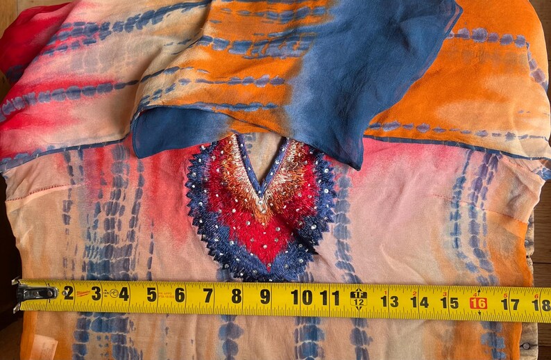 Peut inclure: Une tunique color&eacute;e de style tie-dye avec un d&eacute;collet&eacute; en V. La tunique pr&eacute;sente un m&eacute;lange vibrant de teintes orange, rose et bleu. L'encolure est orn&eacute;e de broderies et de sequins. Le m&egrave;tre ruban indique une largeur d'environ 45 cm.