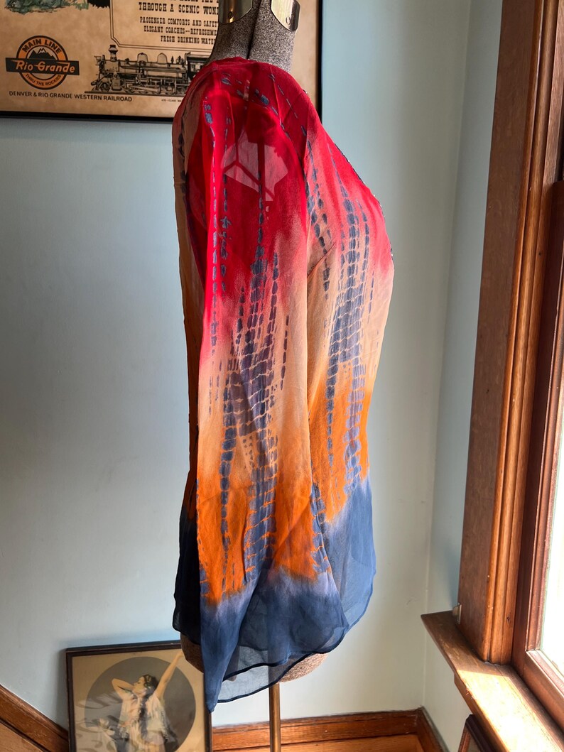 Peut inclure: Un chemisier &agrave; manches longues tie-dye aux tons de rouge, orange et bleu. Le tissu transparent est drap&eacute; sur un mannequin, mettant en valeur le d&eacute;grad&eacute; de couleurs. Une affiche de la compagnie ferroviaire Rio Grande est visible.