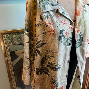 May include: A light-colored button-down shirt with a tropical floral pattern featuring hibiscus flowers and large green leaves. The shirt is measured at approximately 30 inches in length. The shirt is on a mannequin.
